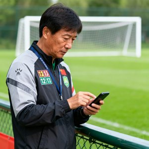 一位穿着运动服装的青训足球教练李明站在训练场边拿着手机查看比分的半身照片